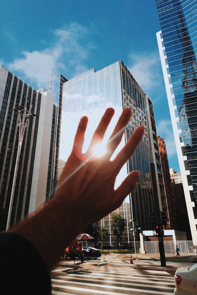 A hand reaches towards modern glass skyscrapers reflecting sunlight in a vibrant urban setting.